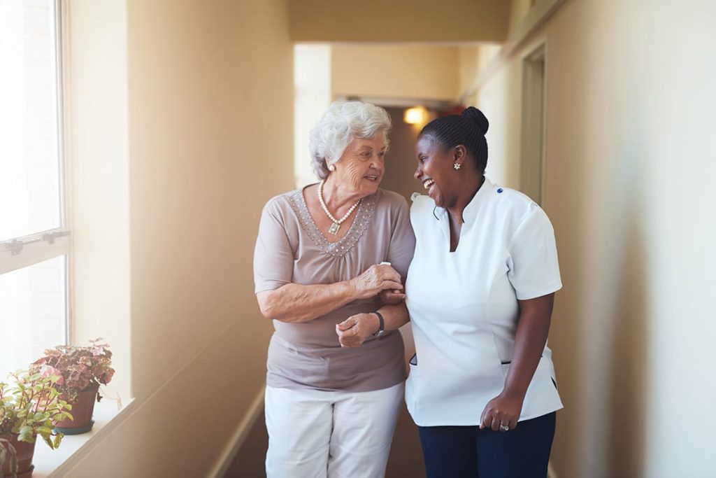 female attendant helping elderly woman walk down the hall as an example of one of the 3 benefits of respite care