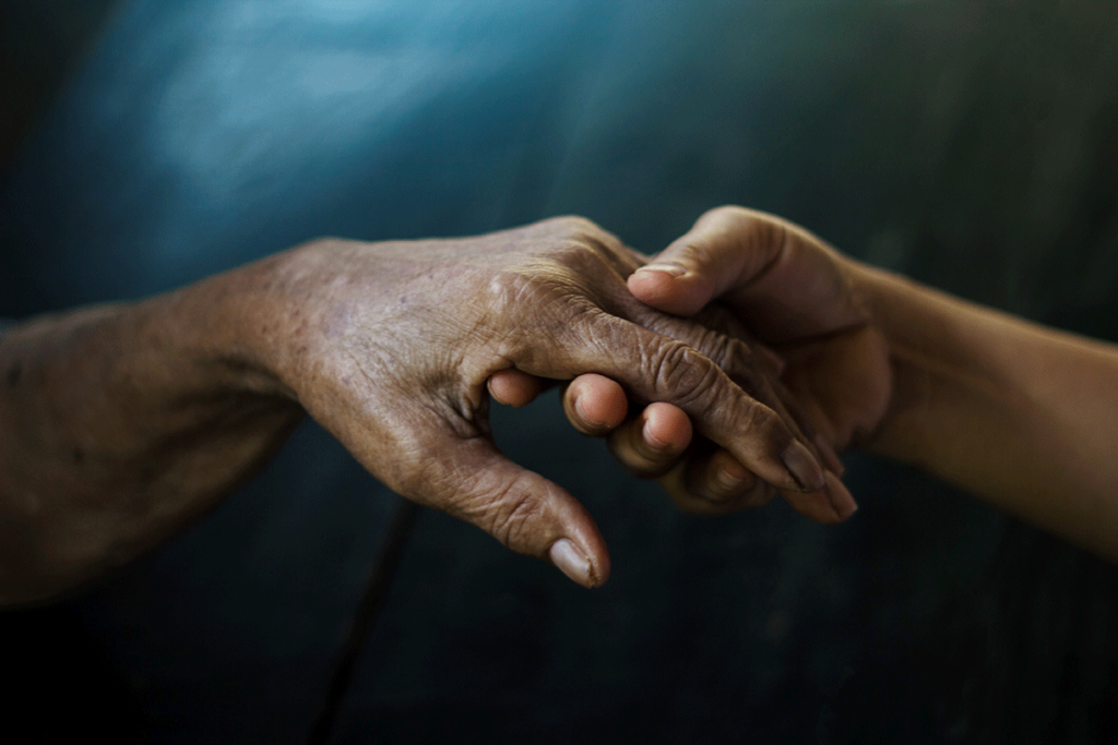 an elderly person in hospice care has their hand held at their bedside