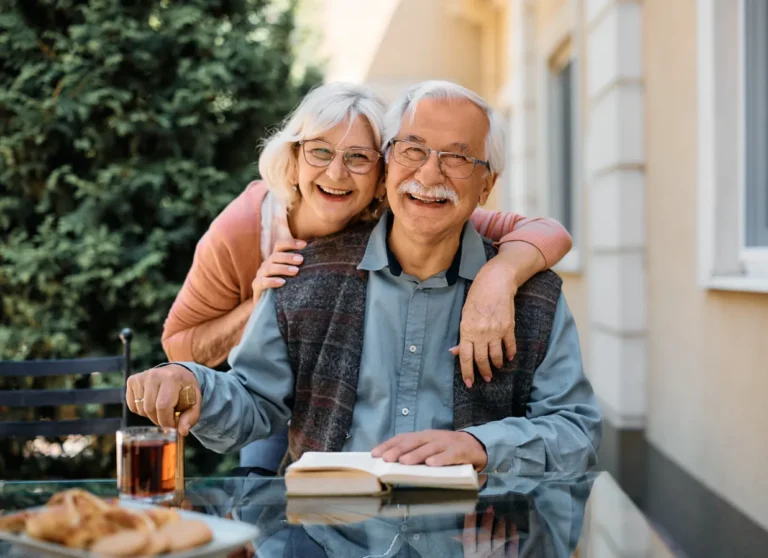 Smiling older couple sits at a glass table outdoors with a book and drink, enjoying each other's company.