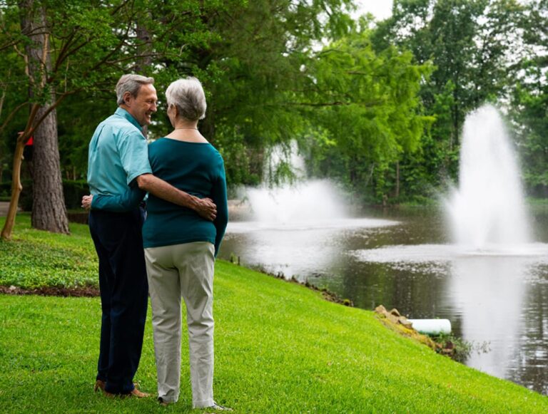 An older couple stands arm-in-arm by a pond with fountains, surrounded by green grass and trees.