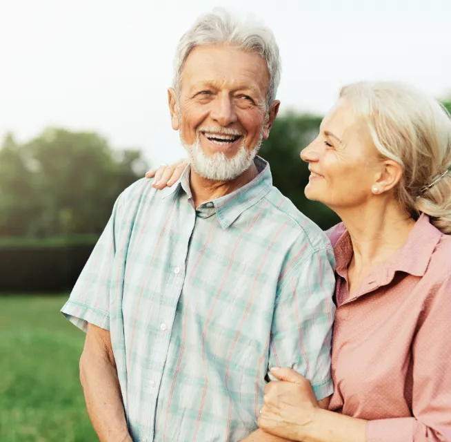 Smiling elderly couple standing outside, the woman holding the man's arm, both looking happy in a green park setting.