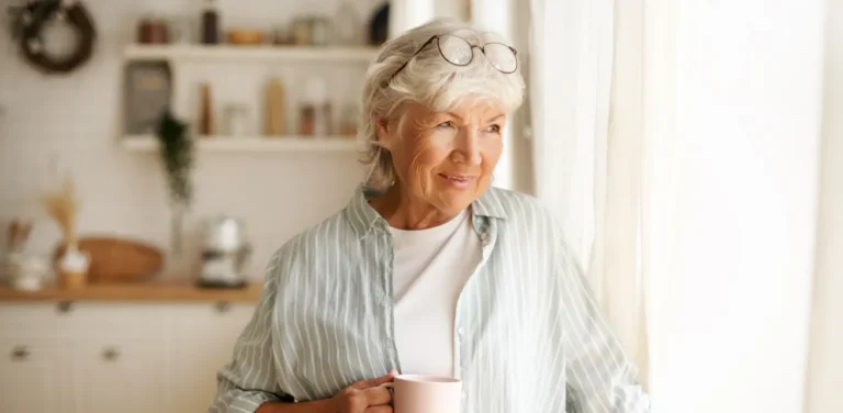 Smiling older woman with gray hair and glasses holds a mug, standing by a bright window in a cozy kitchen.