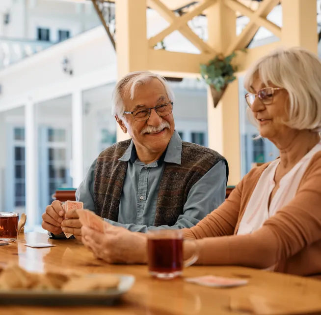Two elderly people sit at a table, smiling and playing cards, with tea and snacks beside them.