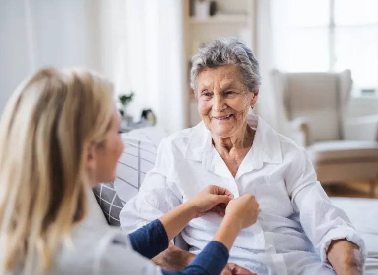 A younger woman helps an elderly woman button her shirt while they both smile in a bright living room.