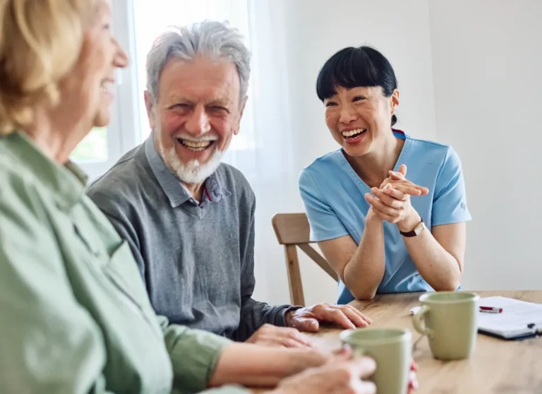 Three people sit at a table, laughing and talking together with mugs and papers in a bright room.