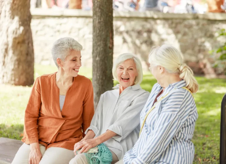 Three older women sit on a park bench, smiling and laughing together on a sunny day.
