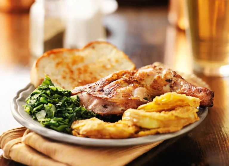 Grilled chicken, sautéed greens, fried okra, and bread on a plate, set on a wooden table with a glass in the background.