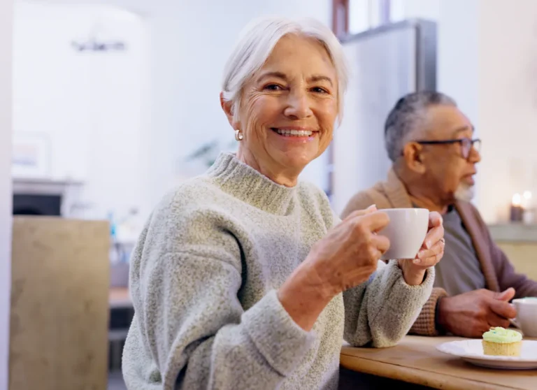 Smiling older woman holding a cup, sitting at a table with a man and a cupcake in a cozy indoor setting.