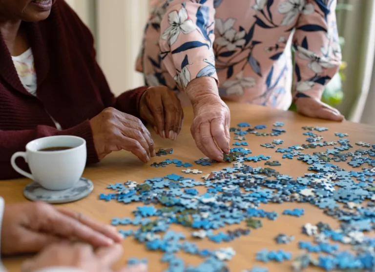 Three people work on a jigsaw puzzle at a table with a cup of coffee nearby. Hands are arranging blue puzzle pieces.