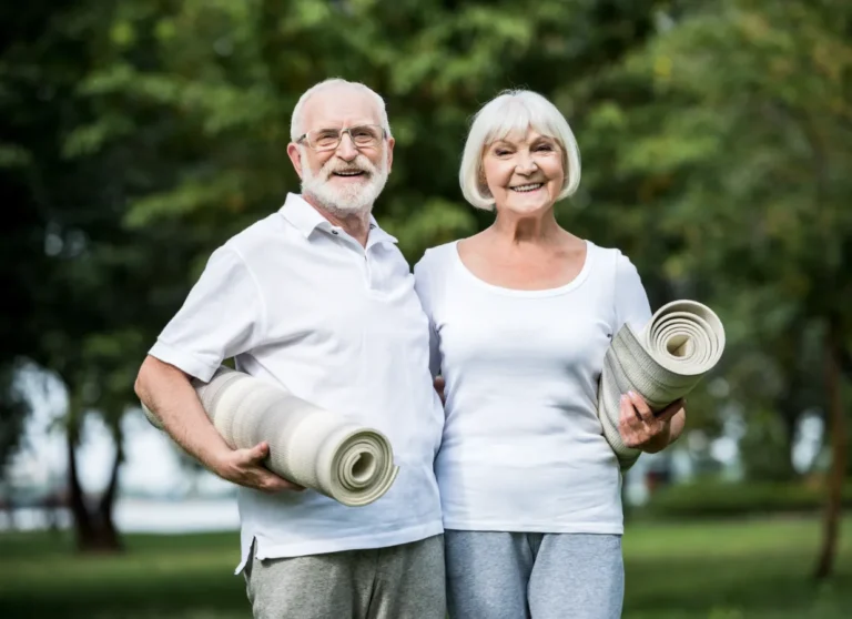 Smiling older couple holding yoga mats outdoors, dressed in casual clothing, with green trees in the background.