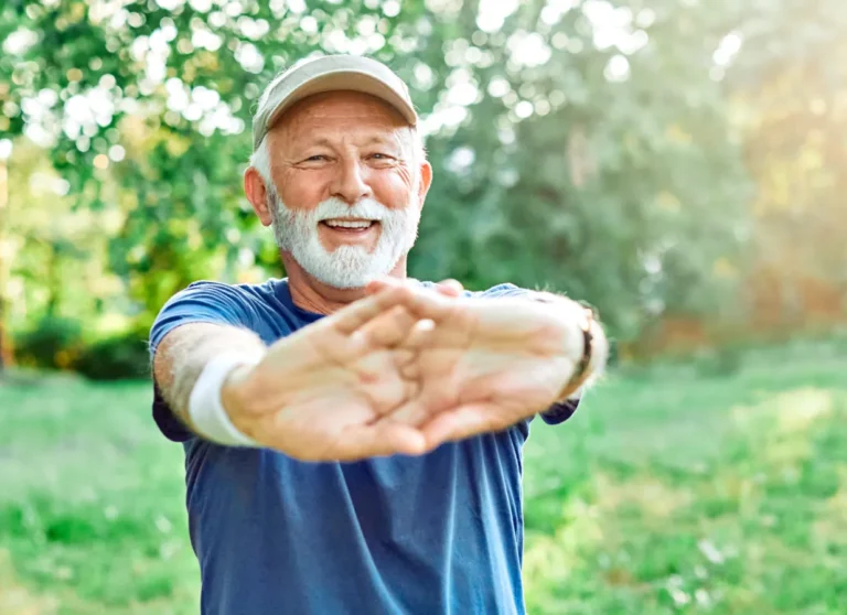 Smiling older man in a cap stretches his arms outside in a sunlit green park.